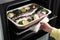 Woman putting baking tray with sea bass fish and vegetables into oven, closeup