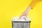 Woman putting shredded paper strips into trash bin on yellow background, closeup