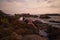 Woman reclines on coastal boulders at dusk, serene sea horizon, waves break nearby, tranquil vacation scene, sunset