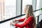 Woman in red coral business suit drinks coffee at a high table near the window. Young blonde businesswoman in the office