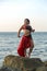 A woman in a red costume for dance with an oriental saber on a belt is posing posing on a rock washed by sea waves.