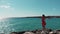 Woman in red dress with hands apart against sun standing on rocky cliff pier enjoying wind and sun. Ocean waves hitting rocky beac