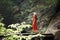 A woman in a red dress and hat poses against a background of rocks covered with moss and ferns. Light from above. The concept of