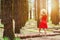 Woman in red dress and hat walks in forest looking at the sun.