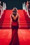 Woman in red dress is walking down red carpeted staircase. Generative AI