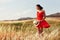 Woman in a red dress walking on the field on a warm summer evening. Yellow grass at sunset, the girl holding a hat in his hands