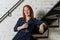 Woman with red hair in dark blue dress holding glass of champagne  sitting on metal ladder loft against white brick wall