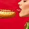 Woman with red lipstick eating spicy hotdog with mustard and chilli over vivid red background. Pepper instead of sausage