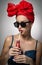 Woman with red turban and sunglasses drinking from a bottle with a straw. Attractive girl portrait holding a bottle, studio shot