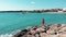 Woman in red walking on rocky beach and raising her hands apart in air looking at blue ocean and sky. Wind is flapping her dress.