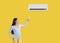 Woman with remote control adjusts comfortable temperature on air conditioner on yellow background.