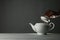 Woman removing lid from ceramic teapot at table against grey background, closeup.