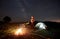 Woman resting at night camping near campfire, tourist tent, bicycle under evening sky full of stars