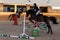 Woman rider on a black steed overcomes the barrier indoors.