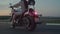 Woman rides a cherry colored motorcycle on highway in evening, then leaves frame, back view. Biker rides motorcycle on