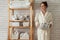 Woman in robe standing and holding white wire basket of folded bed sheets near bathroom linen closet