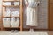 Woman in robe standing and holding white wire basket of folded bed sheets near bathroom linen closet