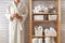 Woman in robe standing and holding white wire basket of folded bed sheets near bathroom linen closet