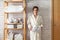 Woman in robe standing and holding white wire basket of folded bed sheets near bathroom linen closet