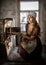 Woman in a rustic dress sitting next to old stove in a ruined abandoned house