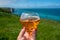 Woman`s hand with glass of apple cider drink and view on  on green grass fields on chalk cliffs of Etretat with blue Atlantic