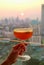 Woman`s hand holding a glass of cocktail with blurry sunset over skyline in background
