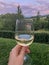 Woman`s hand holding glass of white wine across beautiful mountains landscape and sunset. Vertical. Alcohol. Travel. Green tourism