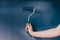 Woman`s hand holding paintbrush roller with dark muted blue tone against matching wall