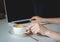 A woman`s hand picks up a white latte Cup in close-up. Coffee stands on a table on a saucer. Hand print text on a laptop