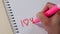 A woman`s hand writes the word I Love You and little heart with a pink marker in a notepad.