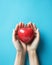 Woman\\\'s hands holding a heart shaped red apple on light azure or blue background