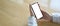 A woman`s hands are holding a smartphone with a white blank screen on the wooden table.