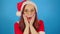 Woman in santa costume opens her mouth in surprise, leaning hands against her cheeks. Female in christmas hat and