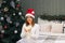 Woman in santa hat with christmas lights in her hands looks at camera in bedroom with xmas tree, festive holiday shine