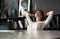 Woman with satisfied expression at desk. Arms up and folded behind her head in modern office.