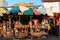 A woman selling coconuts and religious items at a colorful roadside stall under the shade