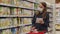 Woman shopper in protecting mask with cart in the store aisle with grocery list during shopping food. Female reads in