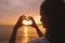 Woman showing shape of a heart with hands on sunset over sea, young woman travel