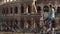 Woman sits on the fence looking at Colosseum in Rome, Italy and swinging her legs. Crowd near the sight. Slow motion.