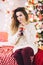 Woman sits near the christmas tree with buttle of cola drink, pretty woman in sweater, holiday atmosphere and garlands