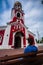 Woman sits on pew near to Church of the Presentation of the Lord in Dmitrov. View from behind on believing girl next to the red