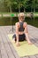 A woman sits on a wooden platform in summer, does yoga on green mat by pond
