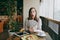 Woman sitting alone in coffee shop at table with bundle of dollars, cash money, cup of cappuccino, cake, relaxing in