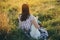 Woman sitting with cute white puppy and looking at sunset in summer meadow. Stylish girl relaxing with her adorable fluffy puppy