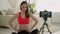 Woman sitting on mat and recording video by smartphone on tripod and doing yoga at home and stretching athletic body