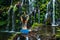 Woman sitting on the rock, practicing yoga. Young woman raising arms with namaste mudra near waterfall. Banyu Wana Amertha