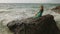 Woman sitting on rock of sea reef stone, stormy cloudy ocean. Blue swimsuit dress tunic. Concept resort coastline