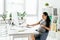 Woman sitting in room with bookcase and flowerpot with plants behind table and working on computer