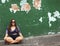 A woman sitting on the sidewalk wearing sunglasses and looking at the camera against an old green colored wall