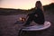 Woman Sitting On Surfboard By Camp Fire On Beach As Sun Sets Behind Her